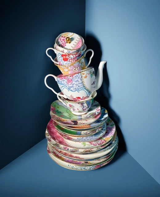 Stack of colorful floral patterned teacups, teapot, and plates in a blue corner setting, creating a whimsical tea party display.