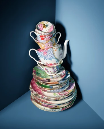 Stack of colorful floral patterned teacups, teapot, and plates in a blue corner setting, creating a whimsical tea party display.