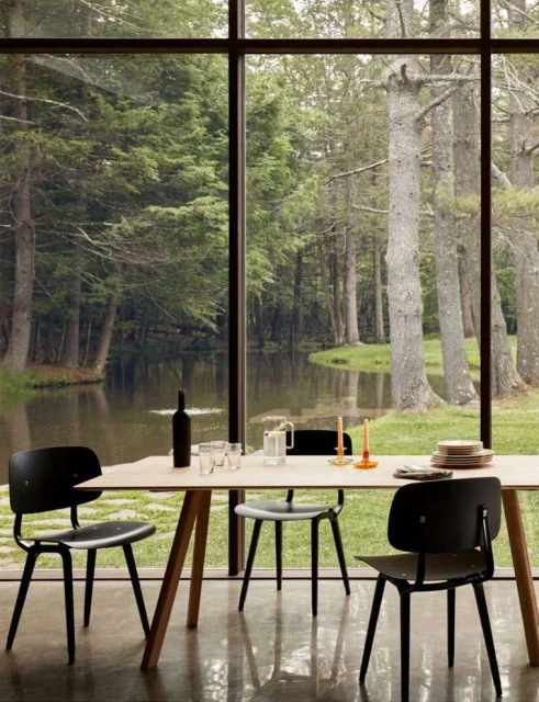 Dining table with black chairs, wine bottle, and glassware set by a large window overlooking a lush forest and a tranquil pond.