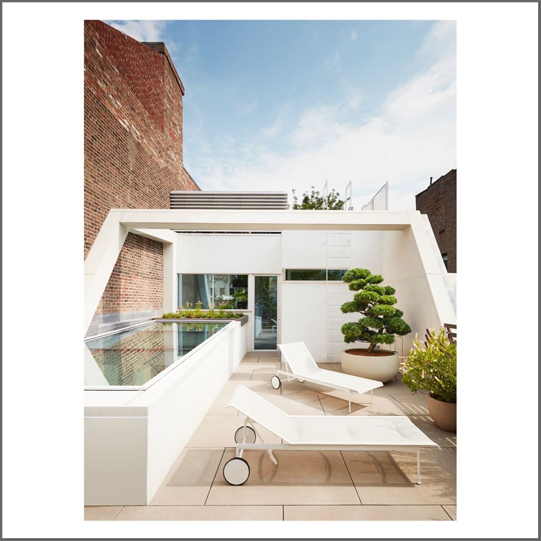 Rooftop patio features modern white lounge chairs, potted bonsai tree, white walls, and brick building under a bright blue sky.