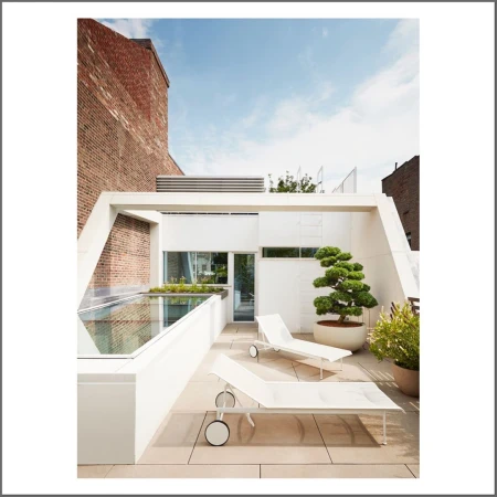 Rooftop patio features modern white lounge chairs, potted bonsai tree, white walls, and brick building under a bright blue sky.