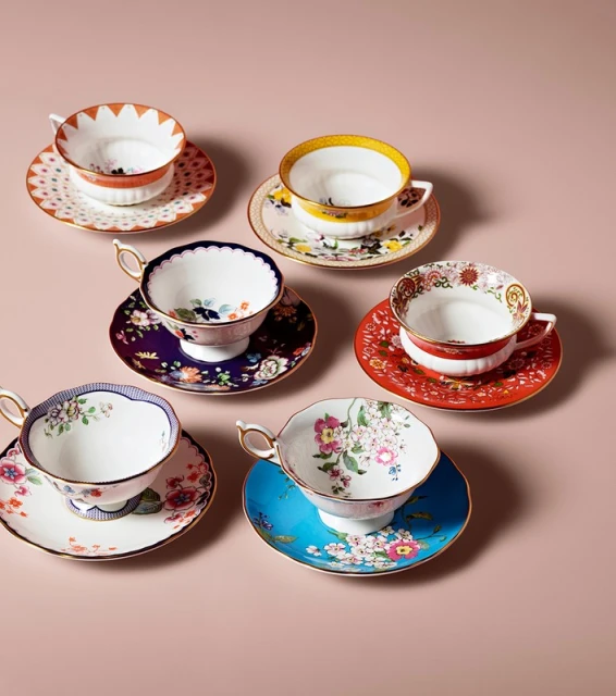 Collection of six antique teacups and saucers with floral and geometric patterns arranged on a light pink surface in a studio setting.