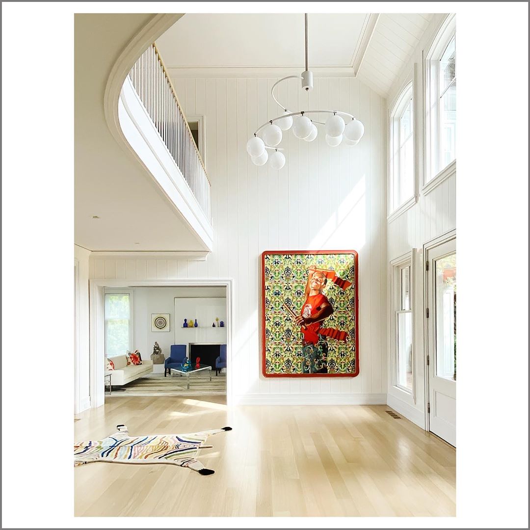 Bright, high-ceilinged interior with a large painting of a person in red tones, modern chandelier, and a view into a living area.