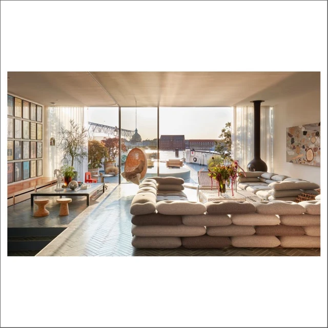 Modern living room interior with large grey sectional sofa, fireplace, coffee table, and view of cityscape through panoramic windows.