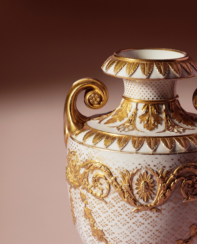 Close-up of a decorative white porcelain vase with ornate gold floral accents, set against a gradient brown background.