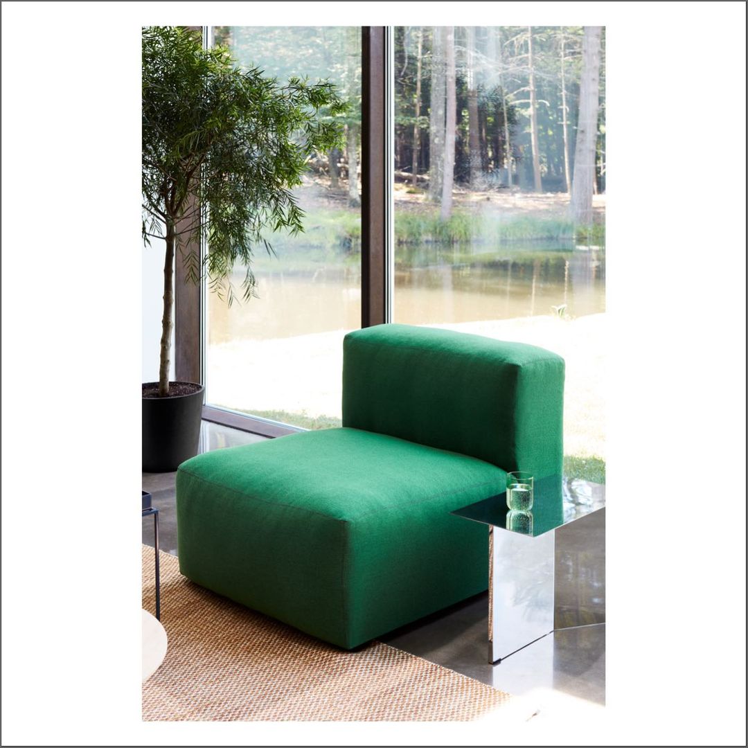 Interior shot of a green upholstered chair with a unique design, complemented by a glass side table, potted plant, and serene lake view through a window.