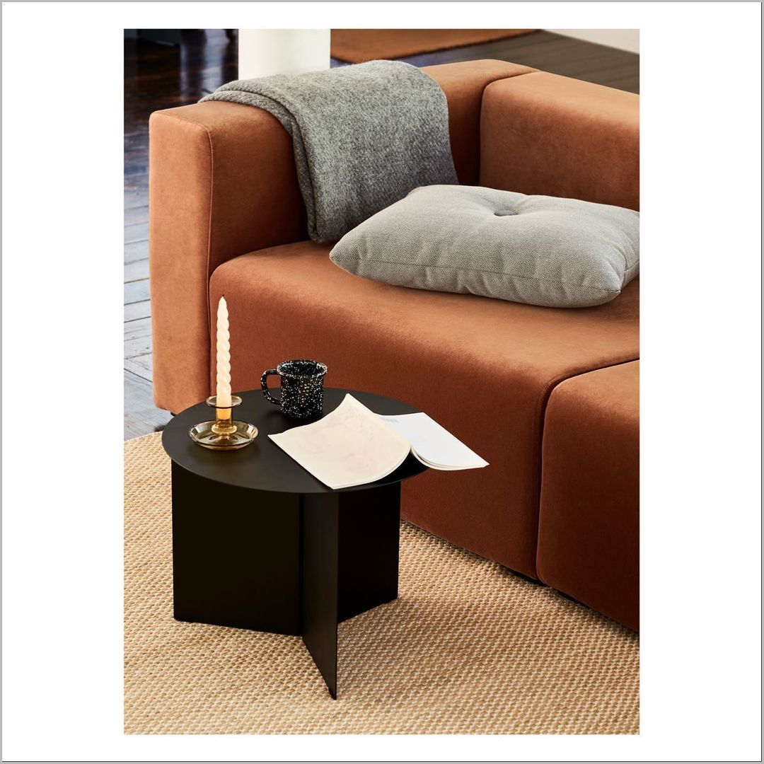 Interior shot of a tan sofa with a gray pillow and blanket, next to a black side table with a candle, mug, and open book.