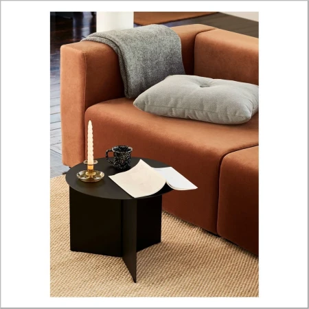Interior shot of a tan sofa with a gray pillow and blanket, next to a black side table with a candle, mug, and open book.