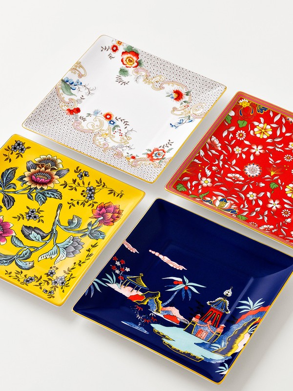 Overhead shot of four square decorative plates with floral and oriental-inspired designs in white, yellow, red, and navy on a white surface.