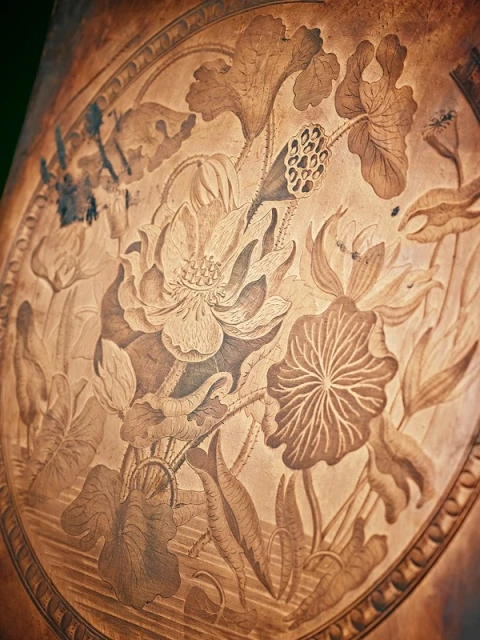 Close-up of a carved wood surface with lotus flowers and leaves; the design features detailed line work and a warm, brown colour palette.