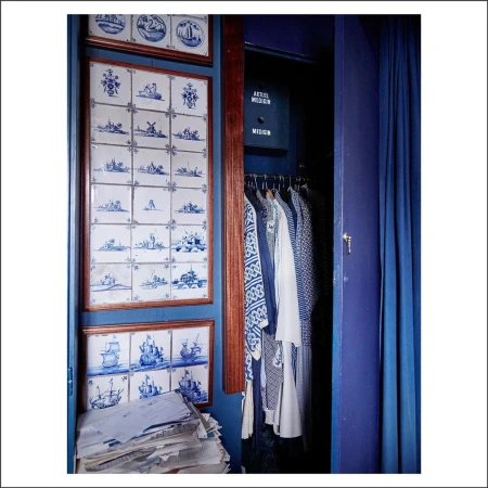 Eclectic interior showcases blue Delft tile art, a clothing-filled closet, and a pile of stacked papers creating a visually intriguing space.
