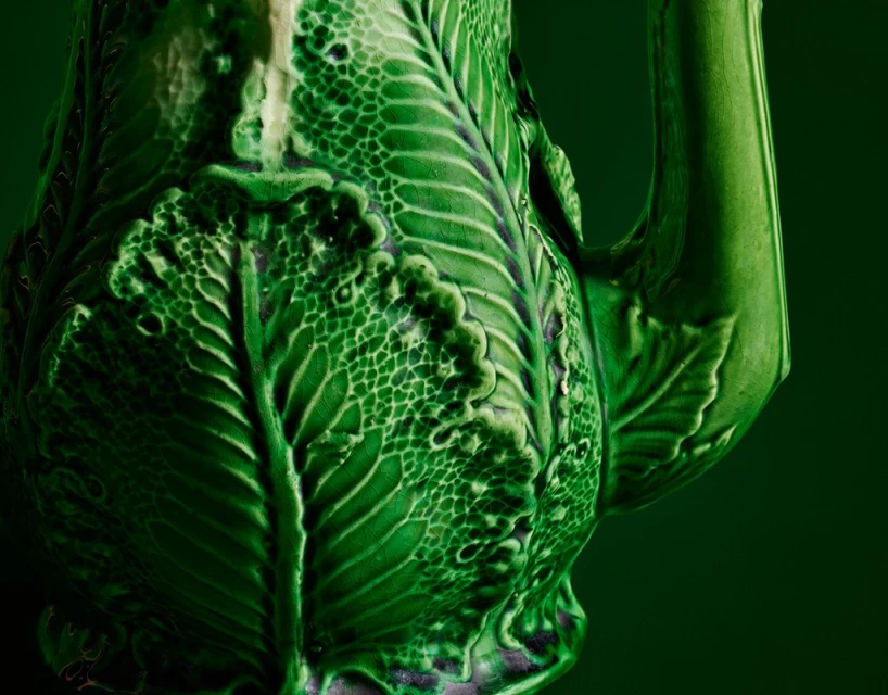Close-up of an ornate, glossy green ceramic pitcher with leaf details on a dark green background showcasing textures and craftsmanship.