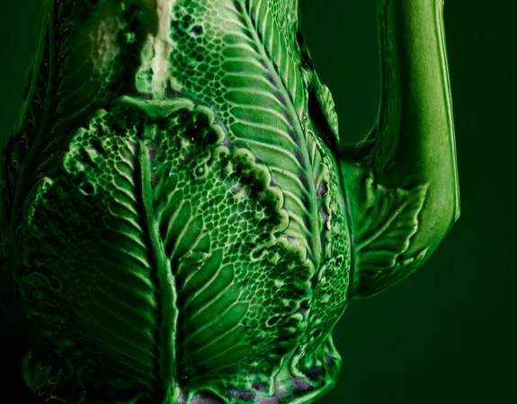 Close-up of an ornate, glossy green ceramic pitcher with leaf details on a dark green background showcasing textures and craftsmanship.