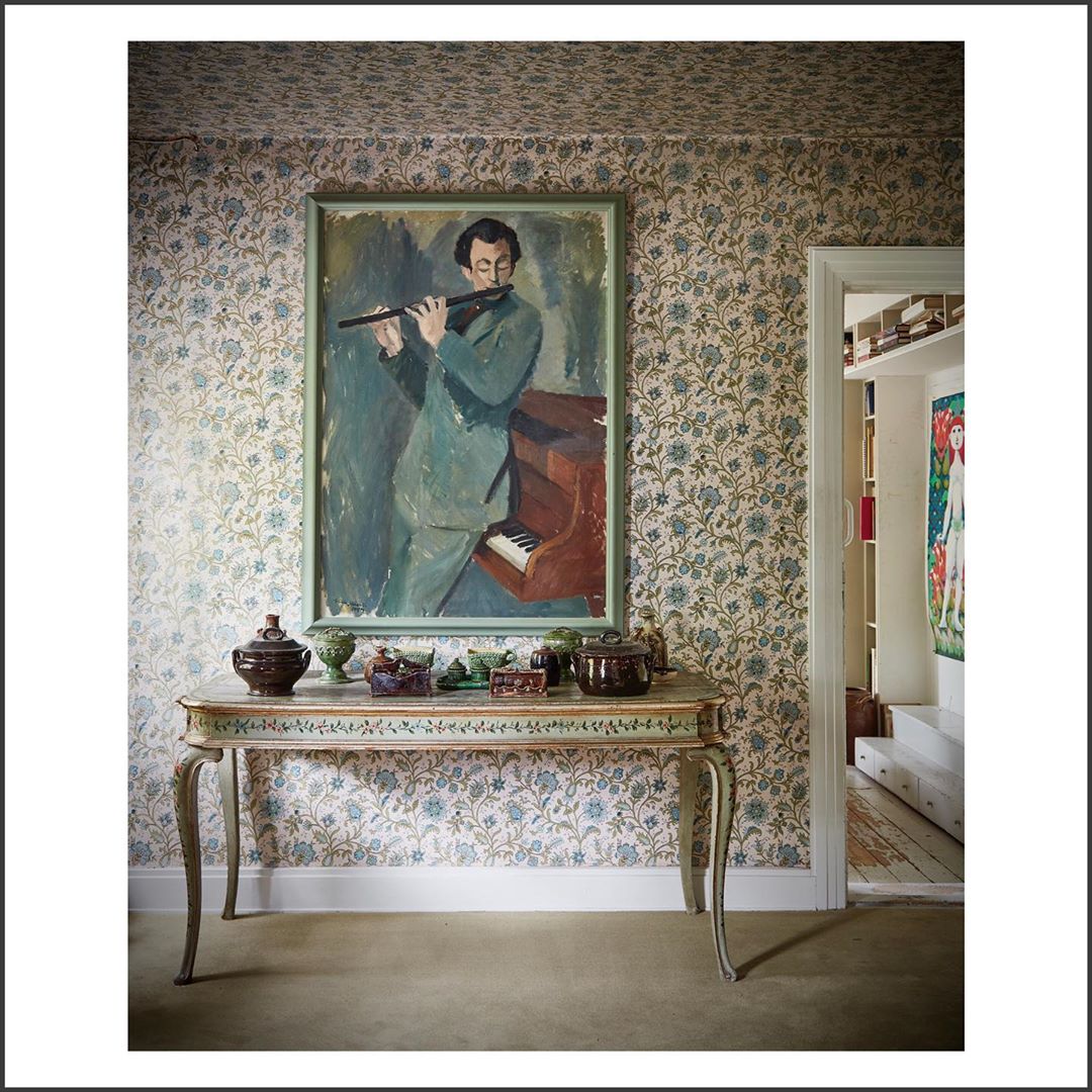 Interior shot featuring a painting of a man playing a flute above a table with decorative objects against floral wallpaper.