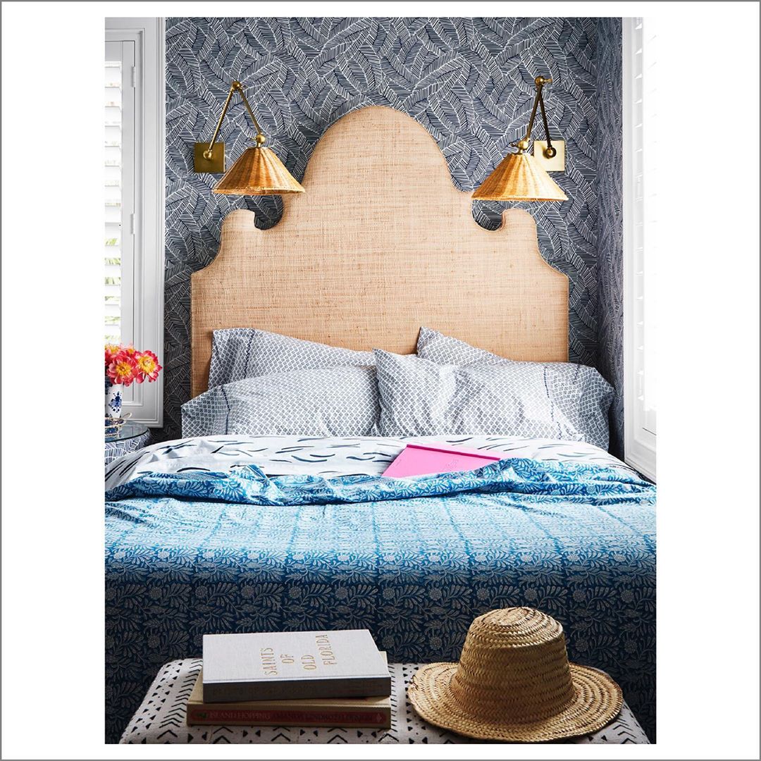 Bedroom interior featuring a bed with a patterned blue quilt, a woven headboard, gold sconces, and leaf-patterned wallpaper.