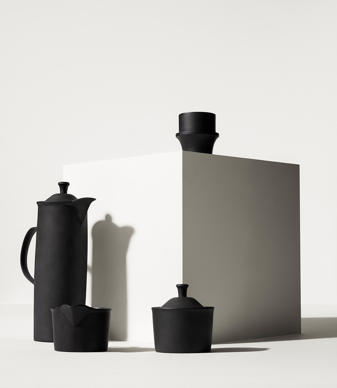 Still life shot of matte black ceramic tea set featuring a tall teapot, creamer, sugar bowl and cup on a white cube in a white studio.