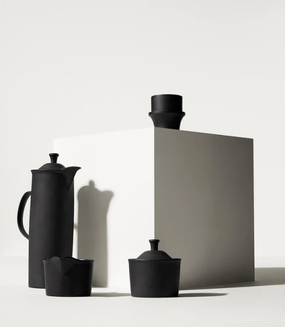 Still life shot of matte black ceramic tea set featuring a tall teapot, creamer, sugar bowl and cup on a white cube in a white studio.