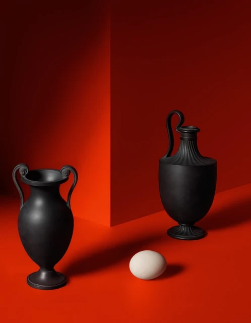 Dramatic studio shot of two matte black decorative vases, one with a white egg, set against a bold gradient red backdrop.
