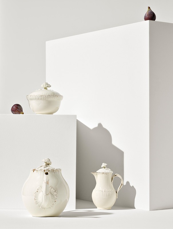 Still life studio shot of ornate creamware teapots and sugar bowl with fresh figs staged on white geometric blocks against white backdrop.