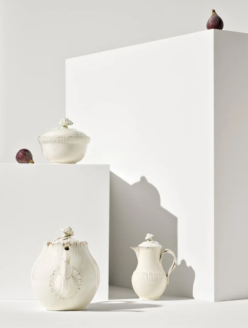 Still life studio shot of ornate creamware teapots and sugar bowl with fresh figs staged on white geometric blocks against white backdrop.