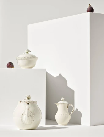 Still life studio shot of ornate creamware teapots and sugar bowl with fresh figs staged on white geometric blocks against white backdrop.