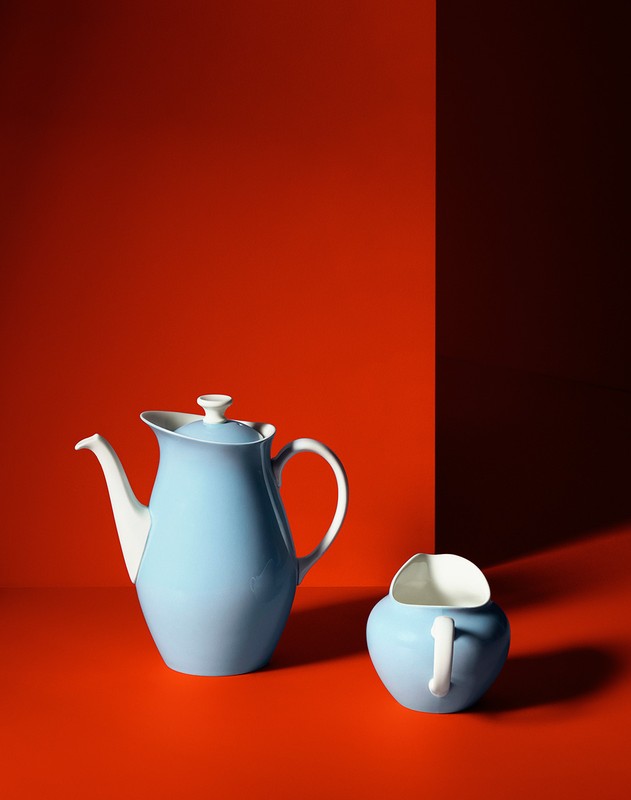 Still life photograph featuring a light blue ceramic tea pot and matching creamer against a vibrant orange background and dark shadow.