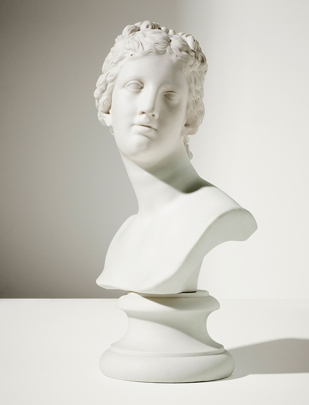 White marble bust of a woman with detailed hair and classical features, set against a plain white background with soft lighting and shadows.