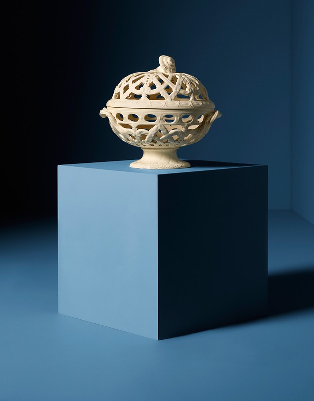 Ornate ceramic pot with a lid on a blue cube against a blue background in a studio setting.