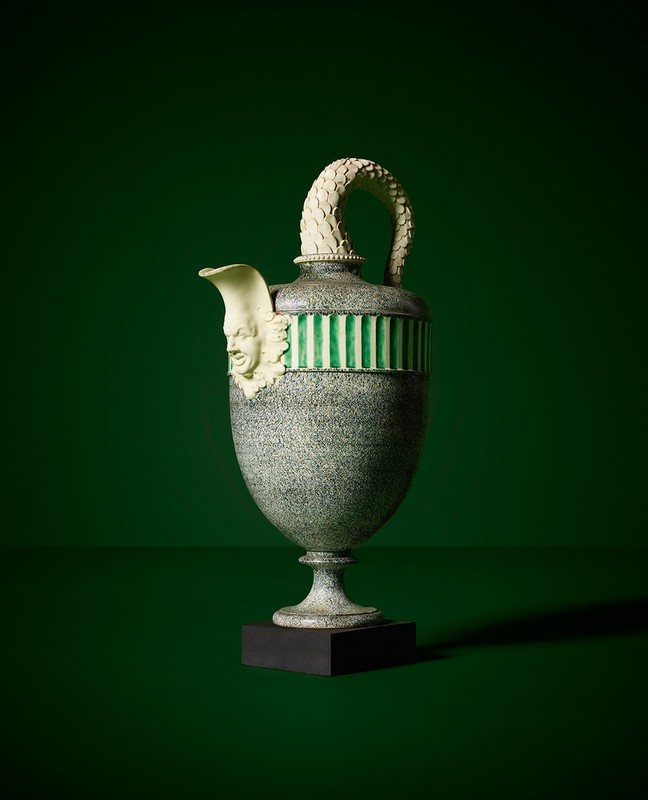 Antique grey ornamental urn with a handle and face detail on a black plinth set against a dark green backdrop.