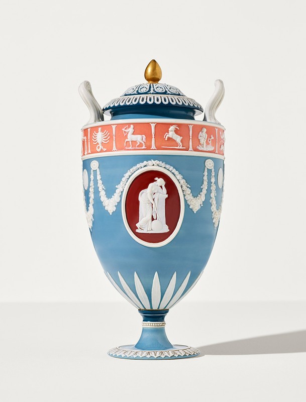 Elegant blue urn with lid, gold finial, relief carvings of human figures and zodiac signs. Ornate ceramic art piece on white background.