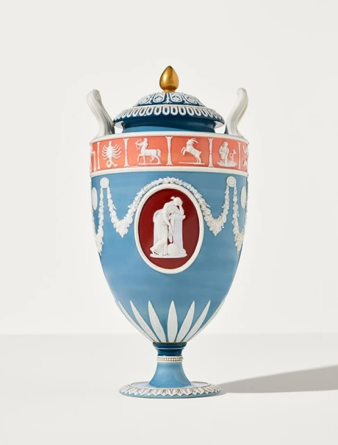 Elegant blue urn with lid, gold finial, relief carvings of human figures and zodiac signs. Ornate ceramic art piece on white background.