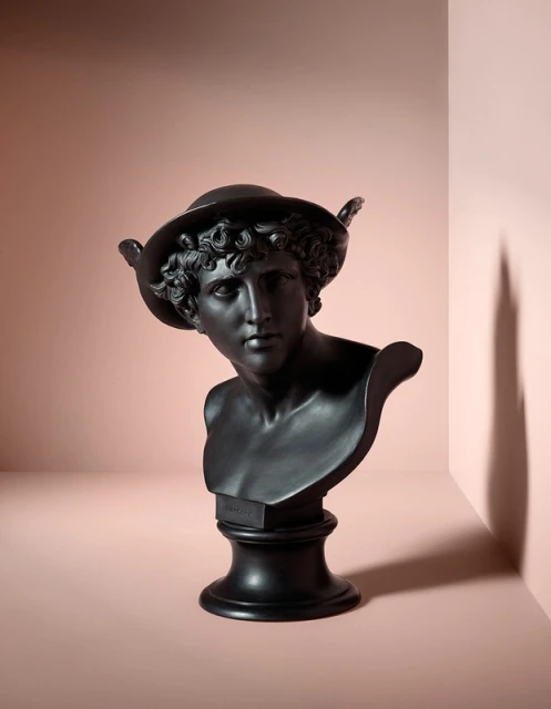 Bust of a statue with a hat and wings in a photo studio on a pink backdrop with shadows.