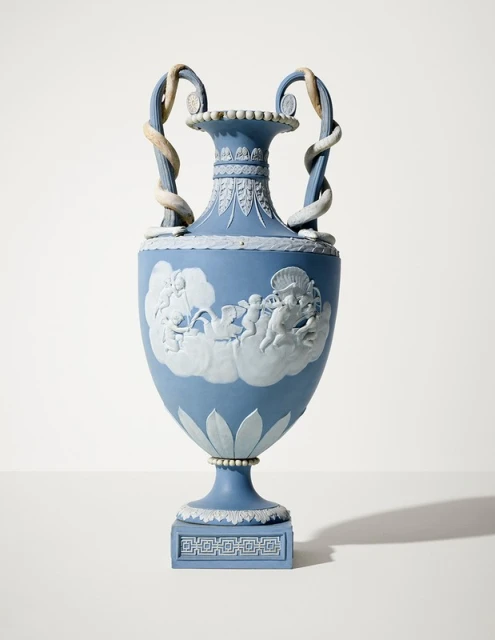 Ornate blue Jasperware vase with white bas-relief figures, serpentine handles, and decorative details against a neutral backdrop.