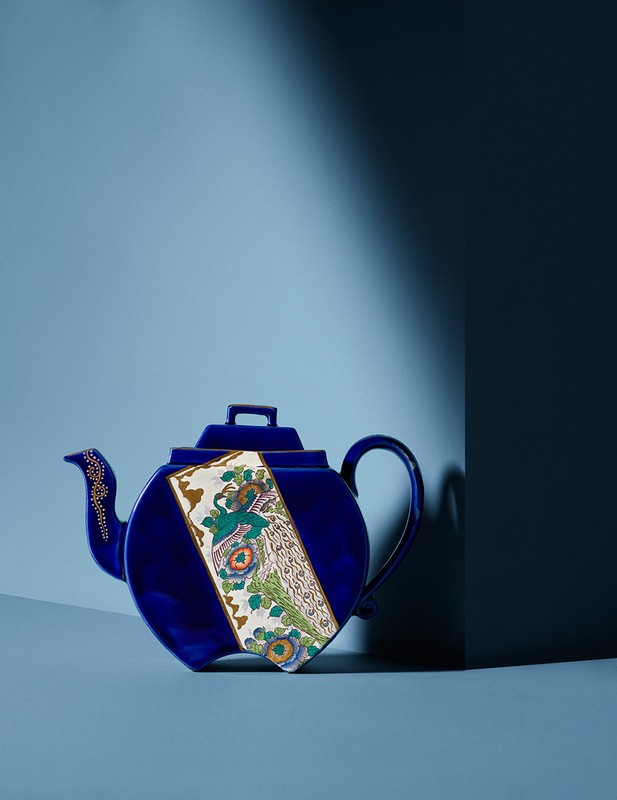 Dark blue, ornate ceramic tea pot sits on a light blue surface in front of a dark blue wall with light and shadow.