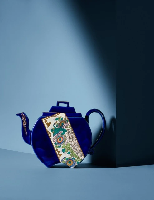 Dark blue, ornate ceramic tea pot sits on a light blue surface in front of a dark blue wall with light and shadow.