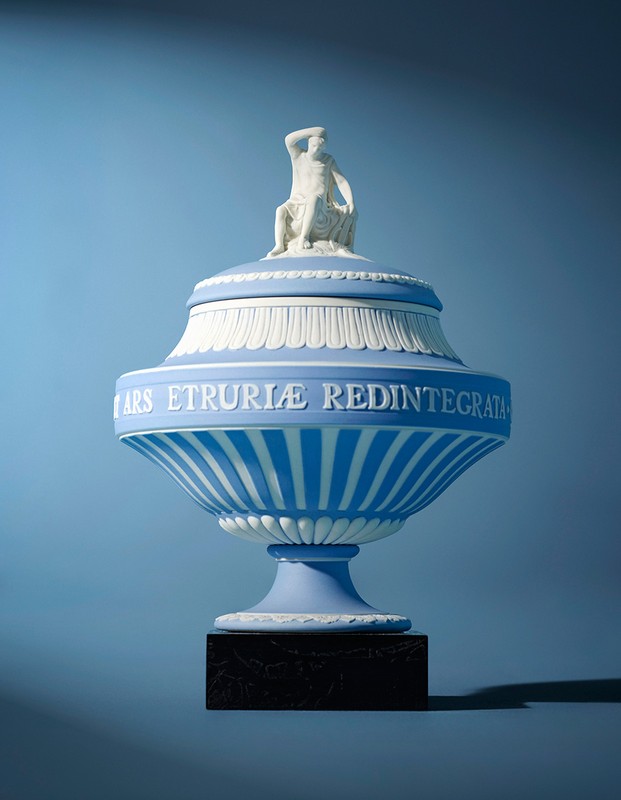 Decorative blue and white Wedgwood urn with sculpted classical figure on the lid against a light blue background in a studio setting.