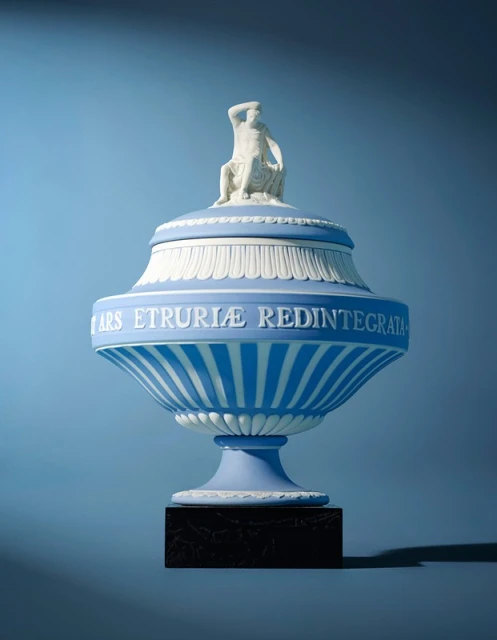 Decorative blue and white Wedgwood urn with sculpted classical figure on the lid against a light blue background in a studio setting.