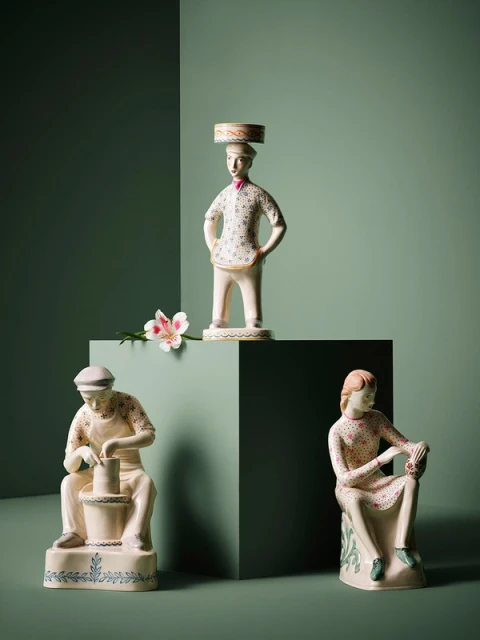 Three vintage ceramic figurines, including a potter, a seated woman, and a man with a hat, arranged on a green background with a flower accent.