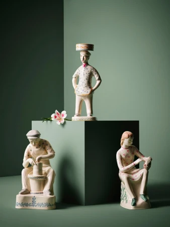 Three vintage ceramic figurines, including a potter, a seated woman, and a man with a hat, arranged on a green background with a flower accent.