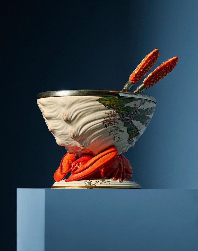 Decorative shell bowl with lobster base, floral details, and salad tongs, set on a blue cube against a gradient blue background.