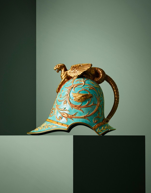 Decorative turquoise helmet with gold accents and a winged lion figure on top, placed against a segmented green background.