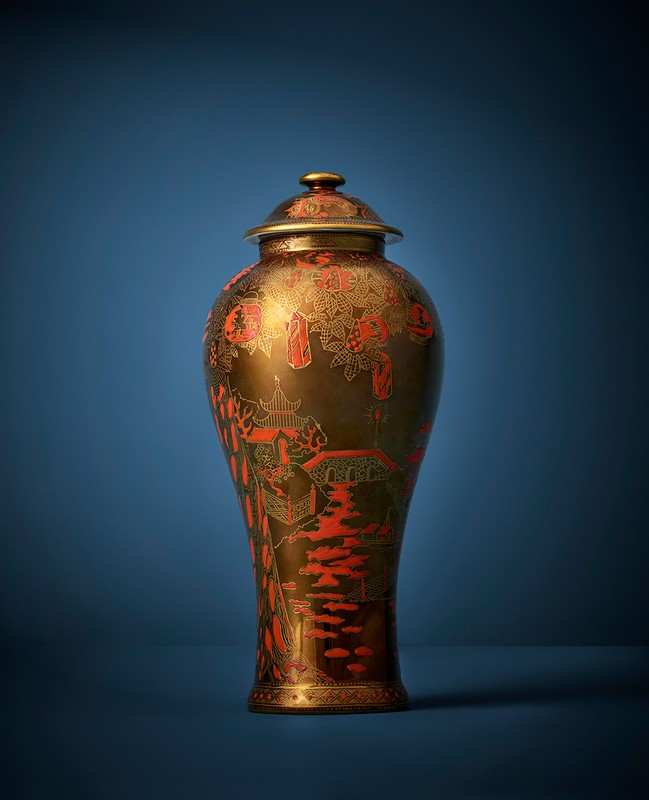 Ornate bronze vase with red detailing and a matching lid on a blue background, standing out with its intricate design and craftsmanship.