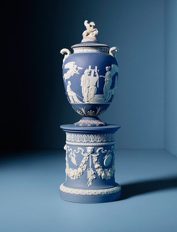 An ornate blue and white Jasperware urn with figures in relief sitting on a matching stand against a graduated blue background.
