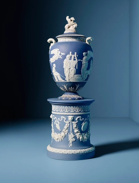 An ornate blue and white Jasperware urn with figures in relief sitting on a matching stand against a graduated blue background.