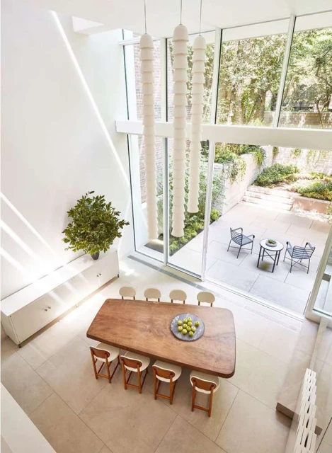 Elevated view of a modern dining room featuring a long wooden table, white chairs, and unique pendant lighting against a bright courtyard backdrop.