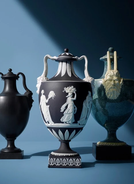 Three decorative urns sit against a blue background, featuring sculpted figures and artistic details in a studio setting.