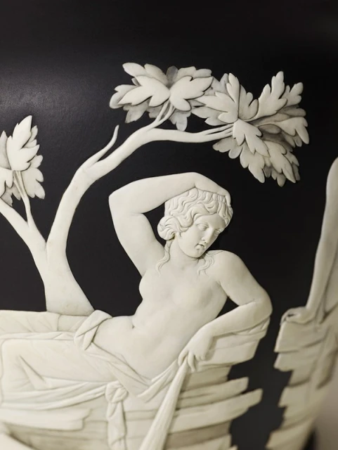 Close-up of a bas-relief sculpture on a black vase featuring a reclining nude woman under a tree. Classic art inspiration.