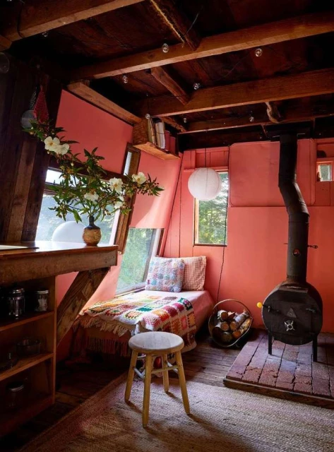 Cozy interior featuring a reading nook with a daybed, wooden beams, a wood-burning stove, floral arrangement, and rustic furniture.