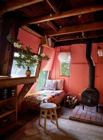 Cozy interior featuring a reading nook with a daybed, wooden beams, a wood-burning stove, floral arrangement, and rustic furniture.