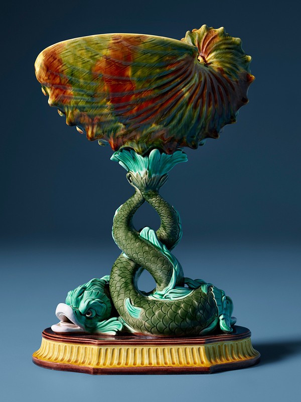 Majolica centerpiece featuring a nautilus shell bowl supported by intertwining fish, set against a soft blue background.